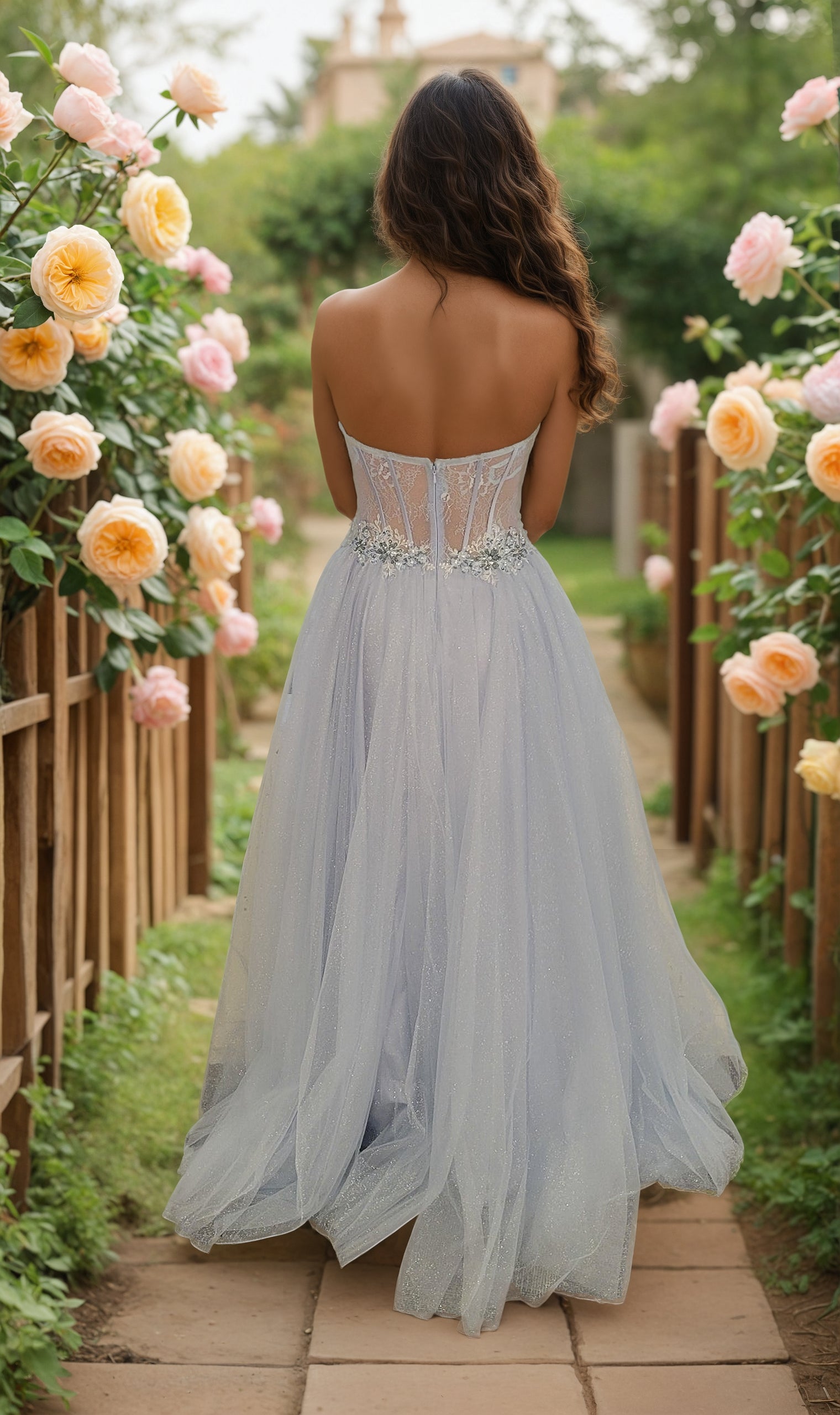 A woman in the Light Blue Prom Ball Gown (Dave and Johnny 12349) with a glitter tulle skirt stands on a garden path lined with blooming yellow and pink roses, facing away from the camera.